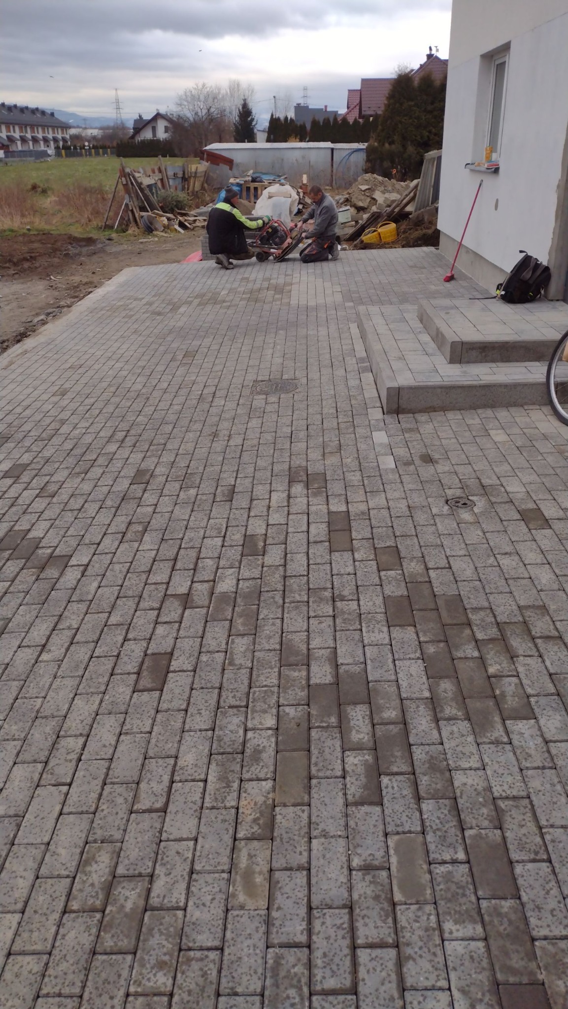 Dwóch pracowników docina kostkę brukową maszyną na nowo ułożonym podjeździe z szarej kostki, widoczne narzędzia, w tle budynek w trakcie wykończenia.