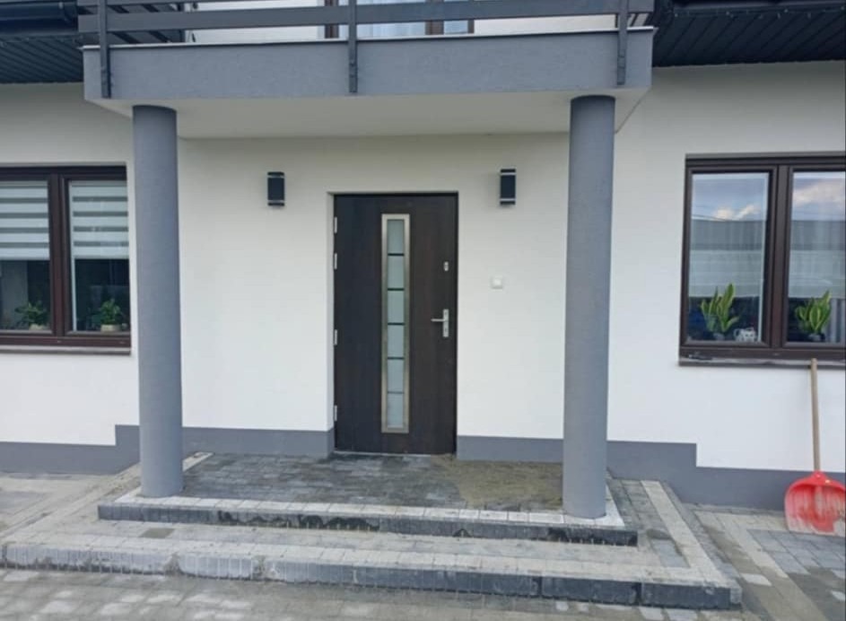 Wejście do domu z ułożoną szarą kostką brukową, widoczne kolumny, drzwi z przeszkleniem, okna z roletami i roślinami, oraz czerwona łopata oparta o ścianę.