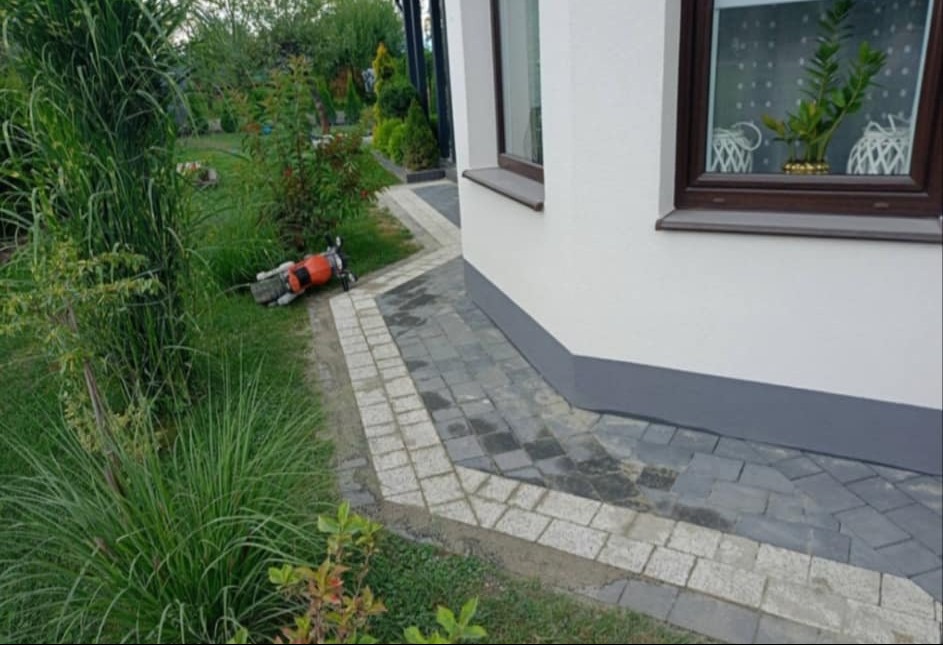 Ułożona ścieżka z kostki brukowej w dwóch odcieniach szarości, obok budynku z brązowymi ramami okiennymi i bujną roślinnością, z pomarańczową zagęszczarką w tle.