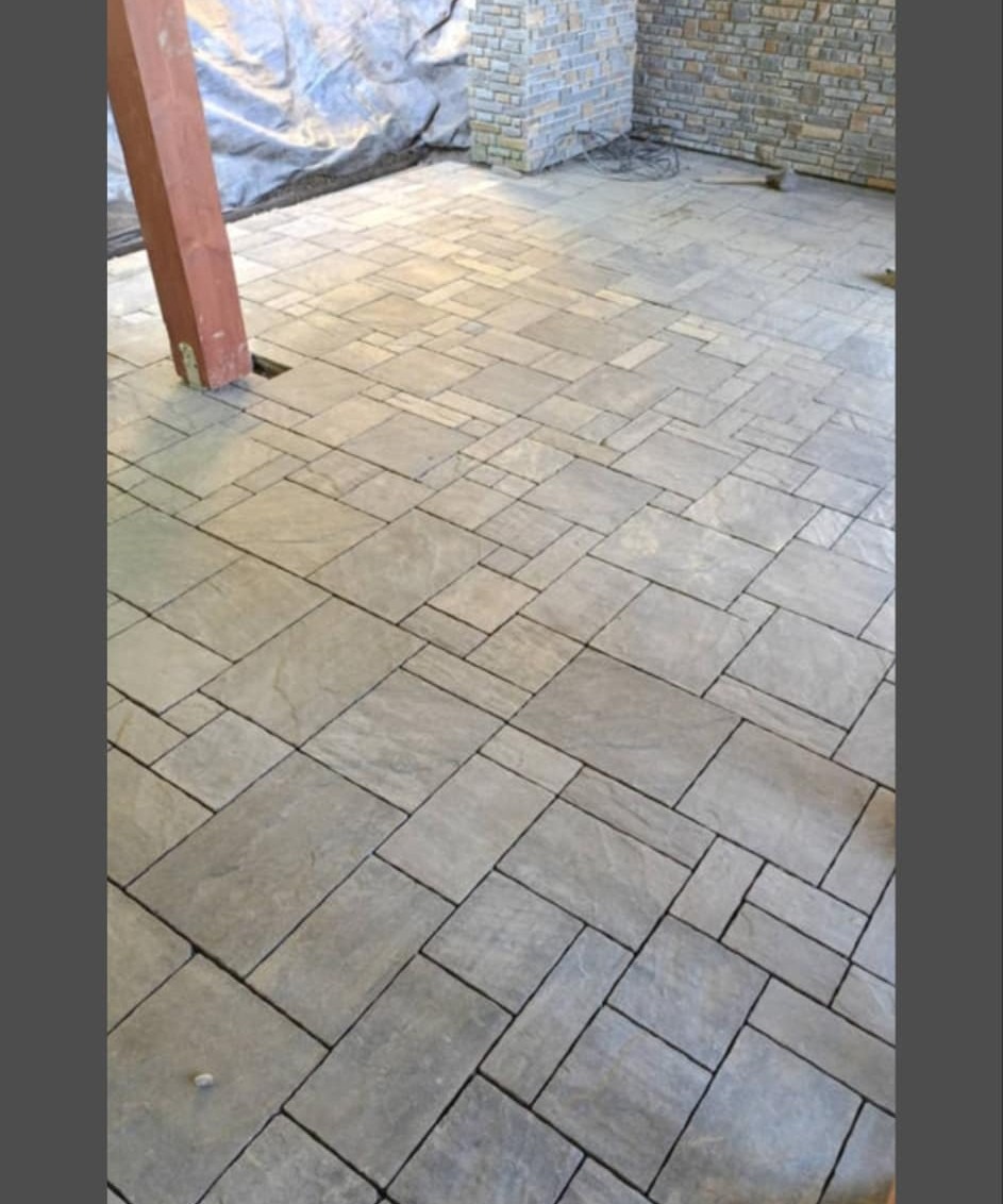 Świeżo ułożona, szara kostka brukowa na tarasie z widocznym drewnianym słupem i kamienną ścianą w tle, częściowo przykrytą folią.