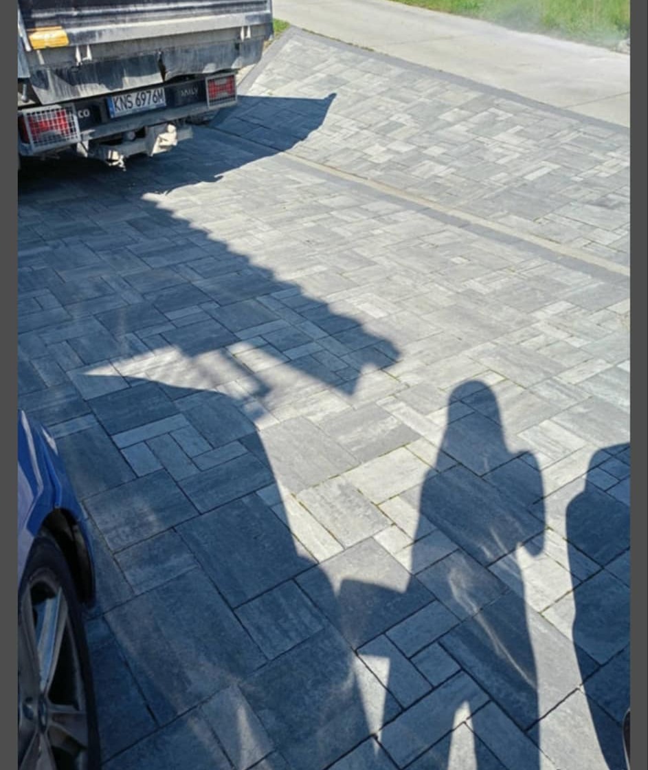 Świeżo ułożona, szara kostka brukowa na podjeździe, z zaparkowanym samochodem dostawczym i rzucanymi cieniami osób, widziana z góry.