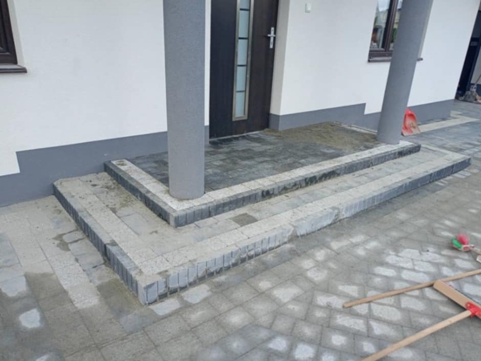 Schody wejściowe do domu wyłożone szarą kostką brukową, z widocznymi śladami mokrego cementu, kolumna podtrzymująca zadaszenie, oraz częściowo ułożony chodnik z kostki.