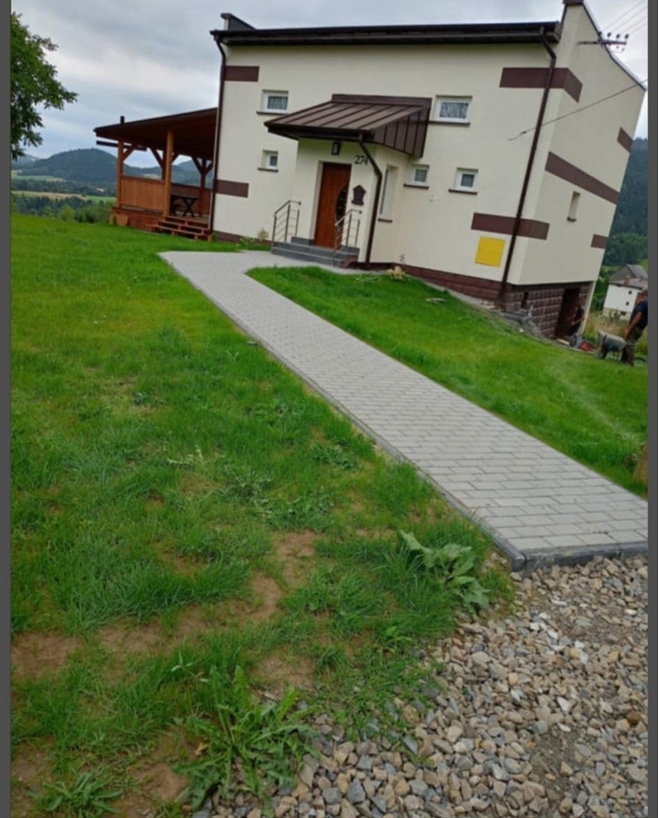 Ułożony chodnik z kostki brukowej prowadzący do domu z brązowymi akcentami, na zboczu wzgórza z widokiem na oddalone góry.