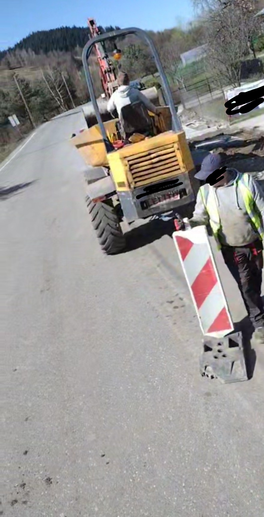 Żółta wywrotka na drodze, pracownik w odblaskowej kamizelce trzyma znak ostrzegawczy, w tle wykop i drzewa.