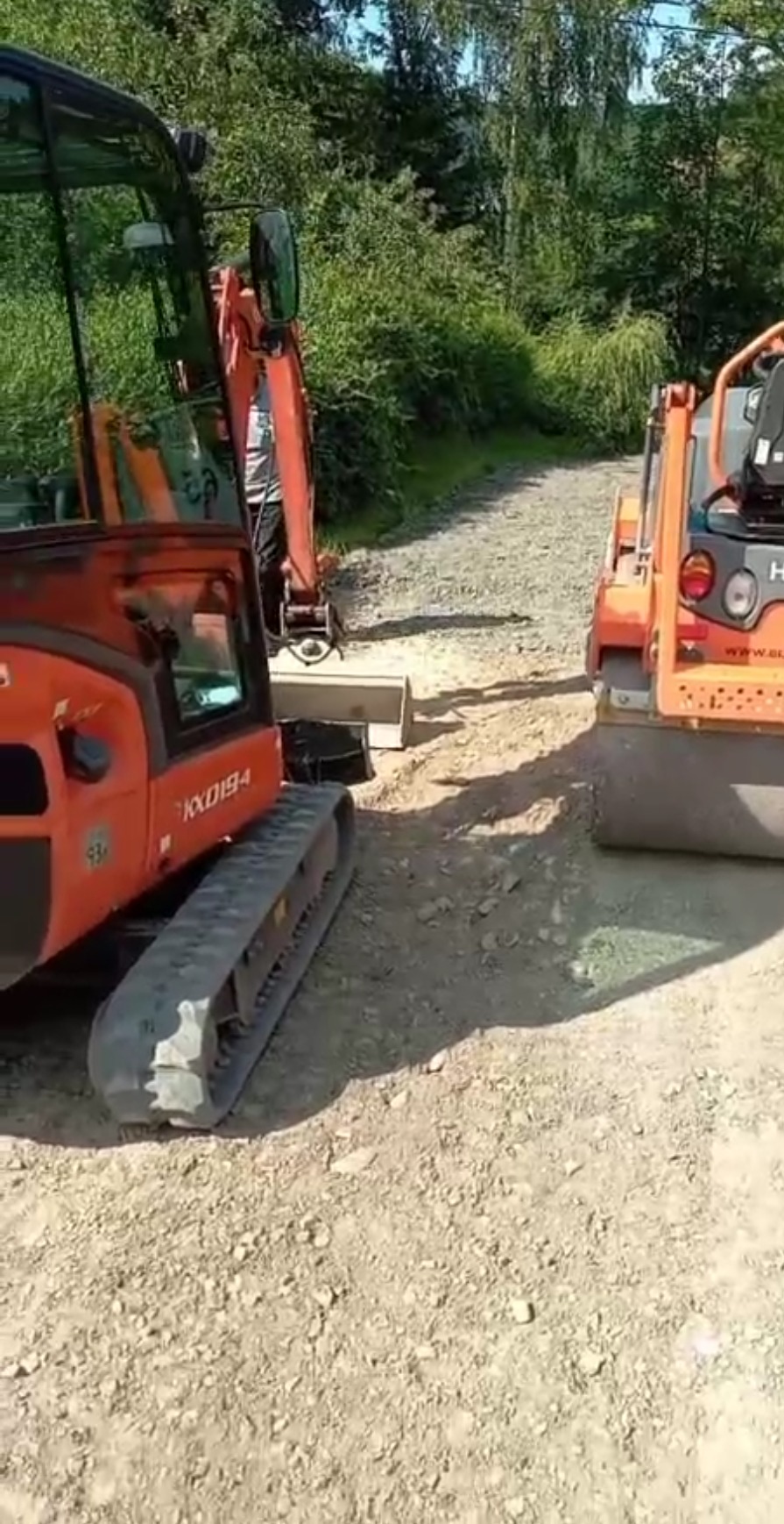 Pomarańczowy walec drogowy i koparka Kubota KX019-4 na żwirowej drodze, w tle zielona roślinność.