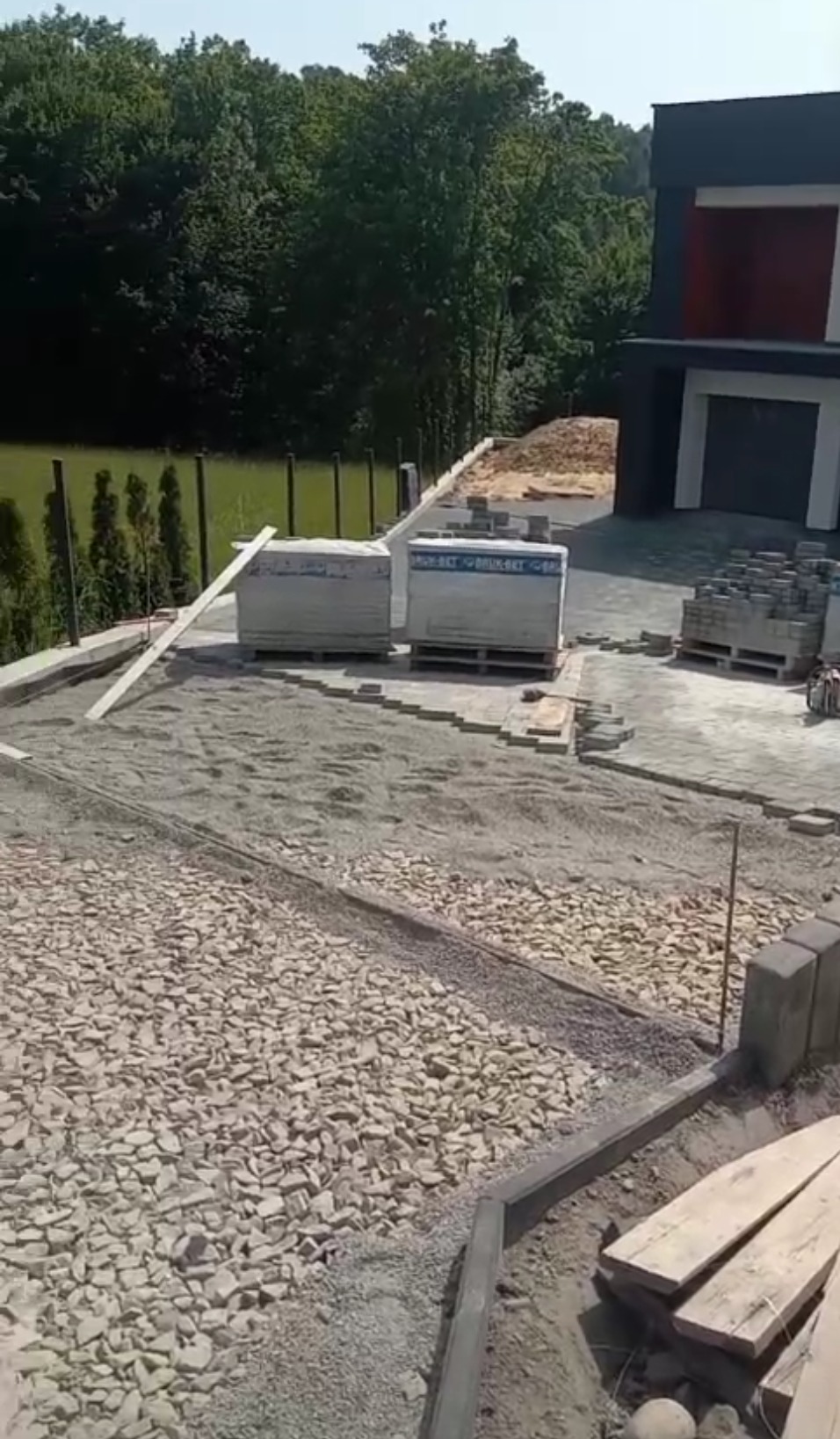 Przygotowanie podłoża pod kostkę brukową: warstwa tłucznia, podsypka piaskowa, obrzeża betonowe, palety z kostką i zapasem materiału, nowoczesny dom w tle.
