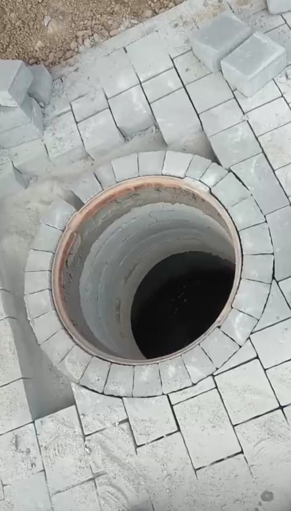 Otwór studni kanalizacyjnej obłożony szarą kostką brukową, częściowo ułożoną, z widocznymi szczelinami wypełnionymi piaskiem, ujęcie z góry.