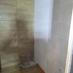 Top Cegła - Wnętrze w trakcie prac: ściany wyłożone płytkami imitującymi drewno i jasnoszarymi, widoczny pion kanalizacyjny wystający z posadzki, przygotowane otwory na armaturę.