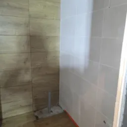 Wnętrze w trakcie prac: ściany wyłożone płytkami imitującymi drewno i jasnoszarymi, widoczny pion kanalizacyjny wystający z posadzki, przygotowane otwory na armaturę.