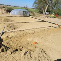 Przygotowanie terenu pod budowę, widoczne wykopane fundamenty, rury kanalizacyjne, poziomica, łopata i szklarnia w tle.