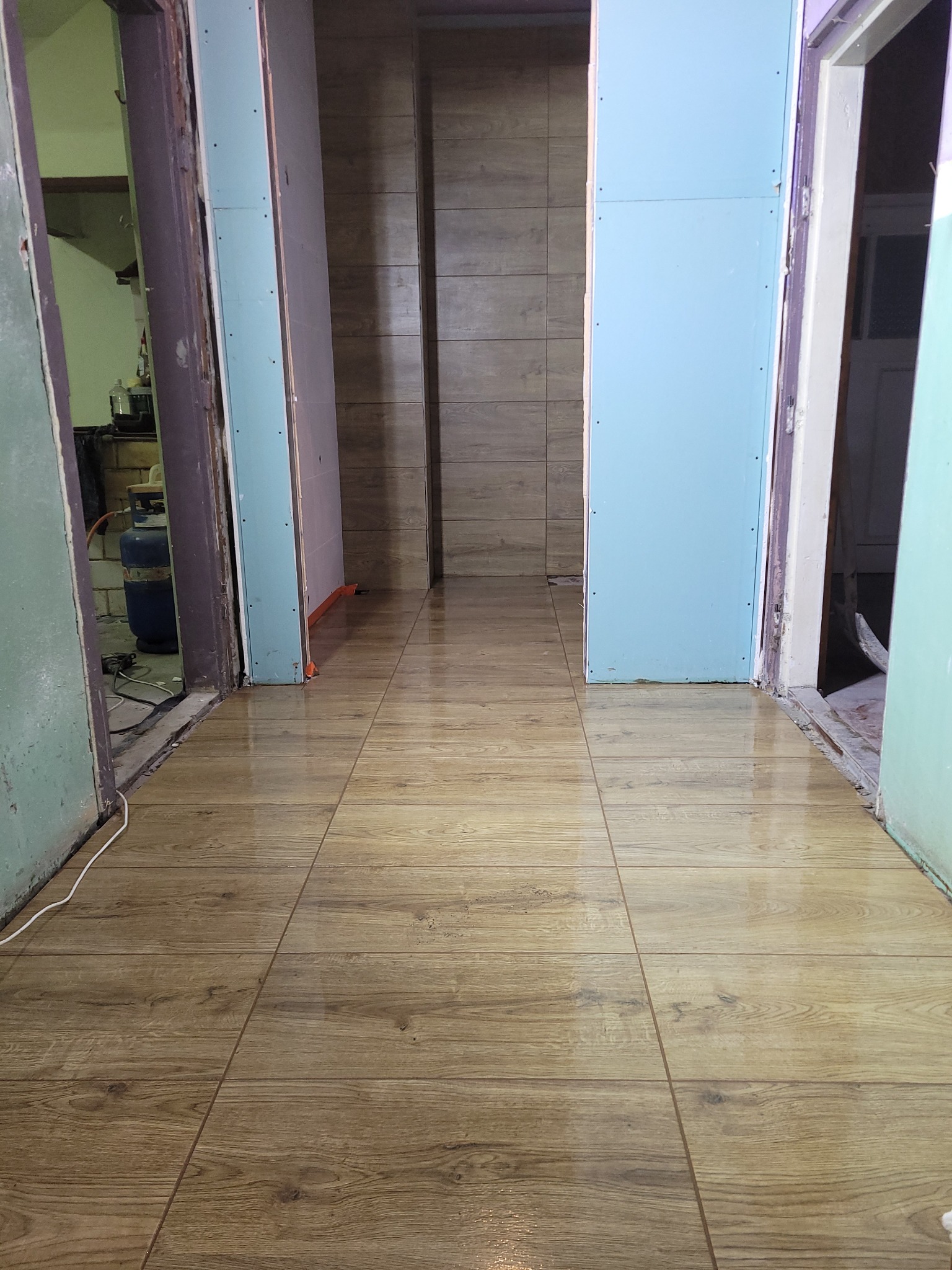 Korytarz w trakcie remontu z nową, połyskującą podłogą z płytek imitujących drewno, widoczne ściany przygotowane pod płyty kartonowo-gipsowe i drzwi wejściowe do pomieszczeń.