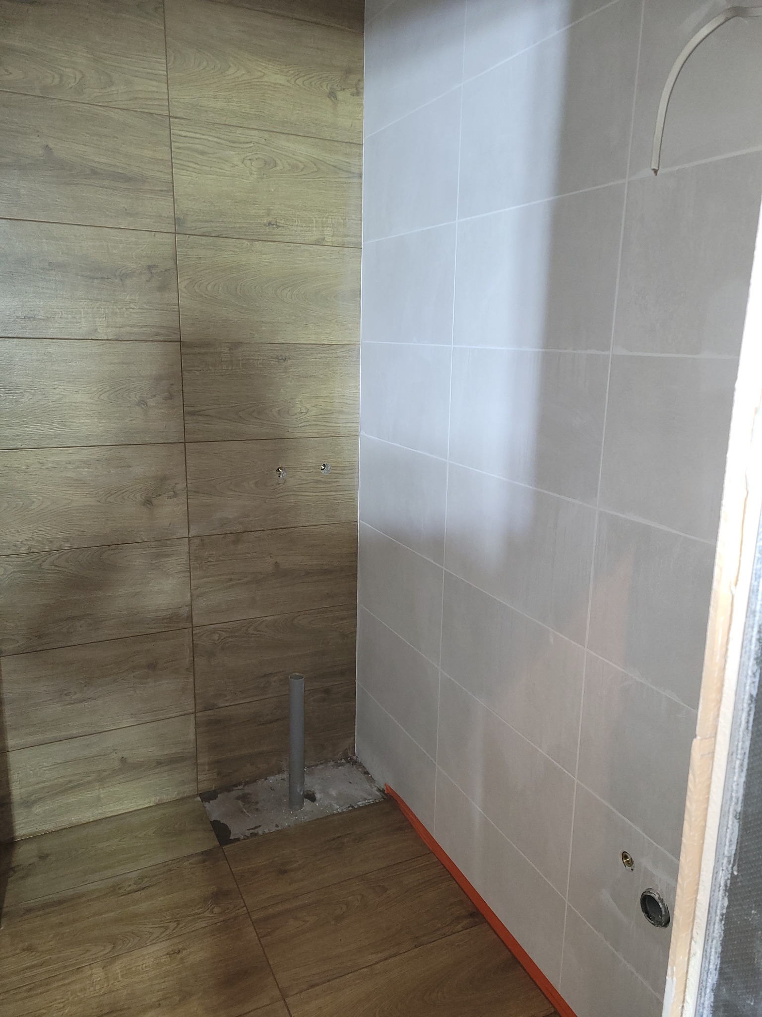 Wnętrze w trakcie prac: ściany wyłożone płytkami imitującymi drewno i jasnoszarymi, widoczny pion kanalizacyjny wystający z posadzki, przygotowane otwory na armaturę.