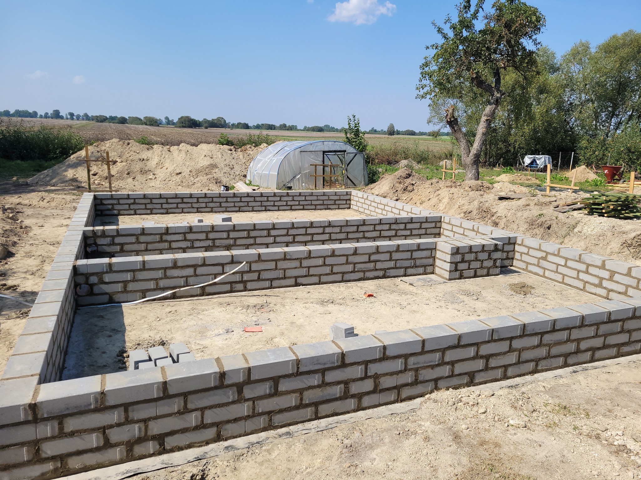 Fundamenty budynku w trakcie budowy, w tle widoczny tunel foliowy i krajobraz polny w słoneczny dzień.