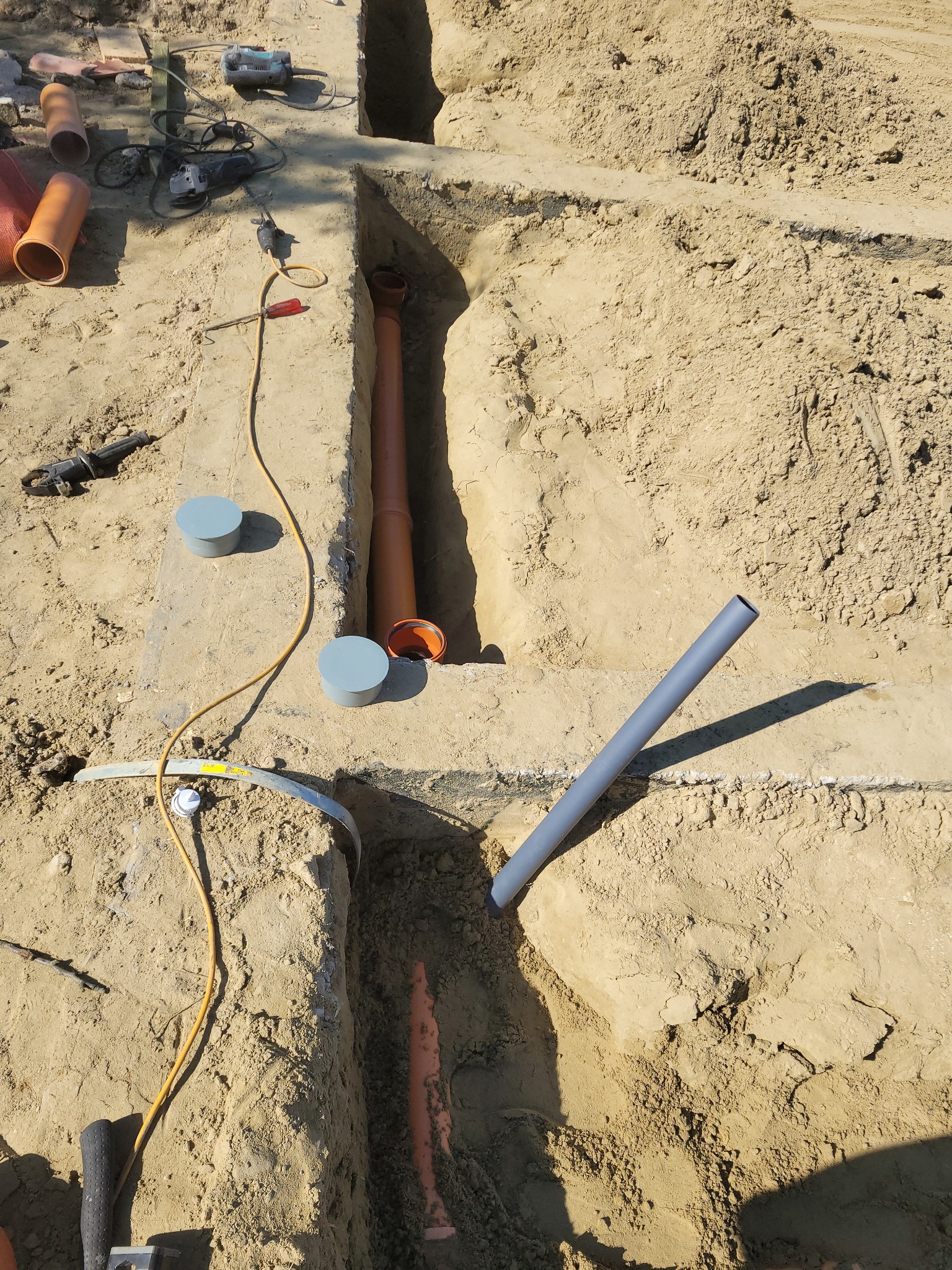 Wykonane wykopy w piasku pod instalacje sanitarne, widoczne rury kanalizacyjne, narzędzia pracy oraz pokrywy inspekcyjne.