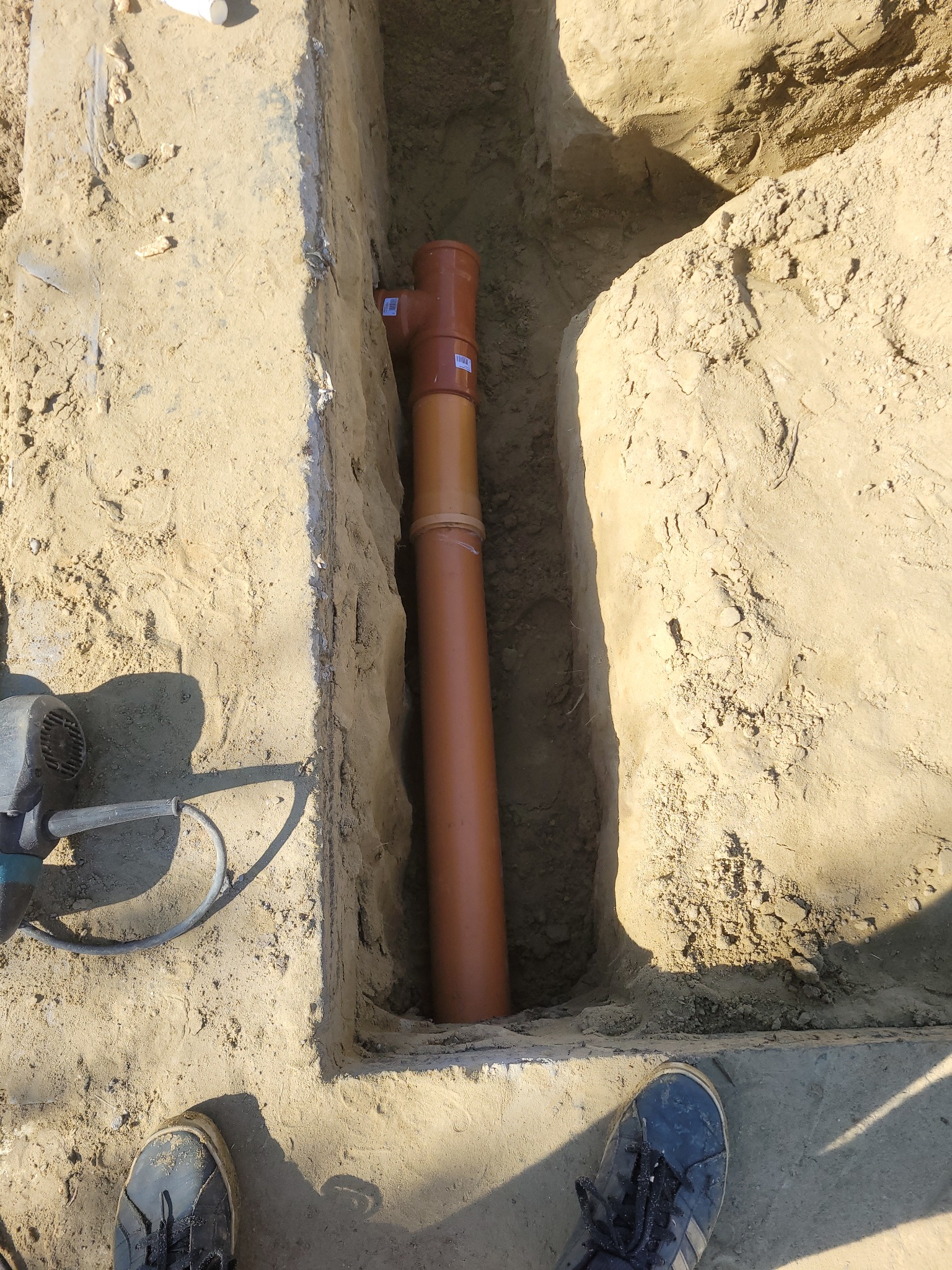 Brązowa rura kanalizacyjna z trójnikiem umieszczona w wykopie w ziemi, widoczny fragment stopy w brudnym bucie sportowym i fragment wiertarki udarowej.