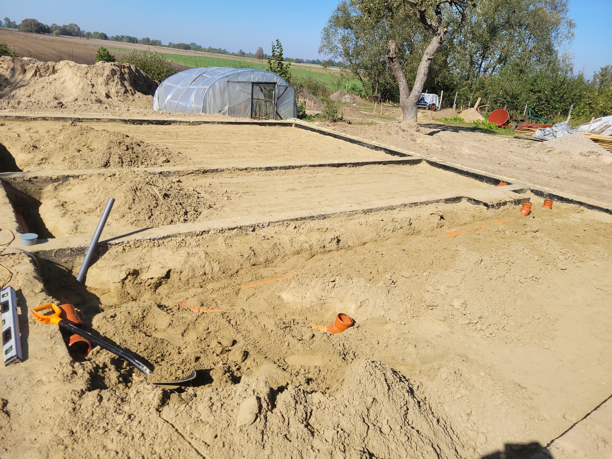 Przygotowanie terenu pod budowę, widoczne wykopane fundamenty, rury kanalizacyjne, poziomica, łopata i szklarnia w tle.