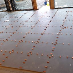 Gresmaster - Układanie szarych płytek podłogowych z pomarańczowymi klipsami poziomującymi i cienkimi czarnymi liniami fug, widoczny częściowo pracownik i lampa budowlana