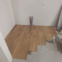 Gresmaster - Układanie podłogi z płytek imitujących drewno w trakcie prac, widoczne rury instalacyjne na ścianie i częściowo położone płytki.