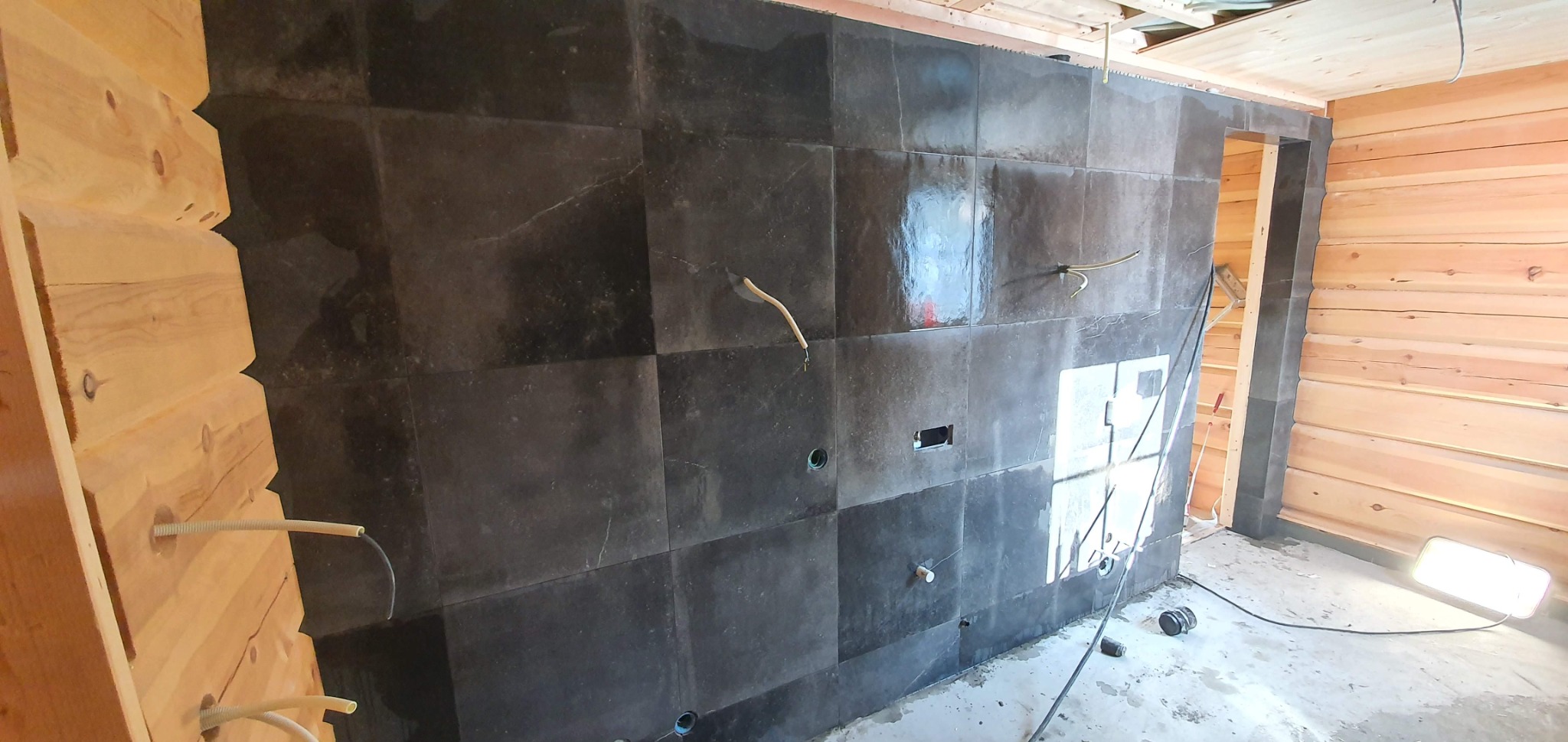 Ściana z ciemnych, kwadratowych płytek w trakcie układania w drewnianym wnętrzu. Widoczne wystające kable i otwory na instalacje.