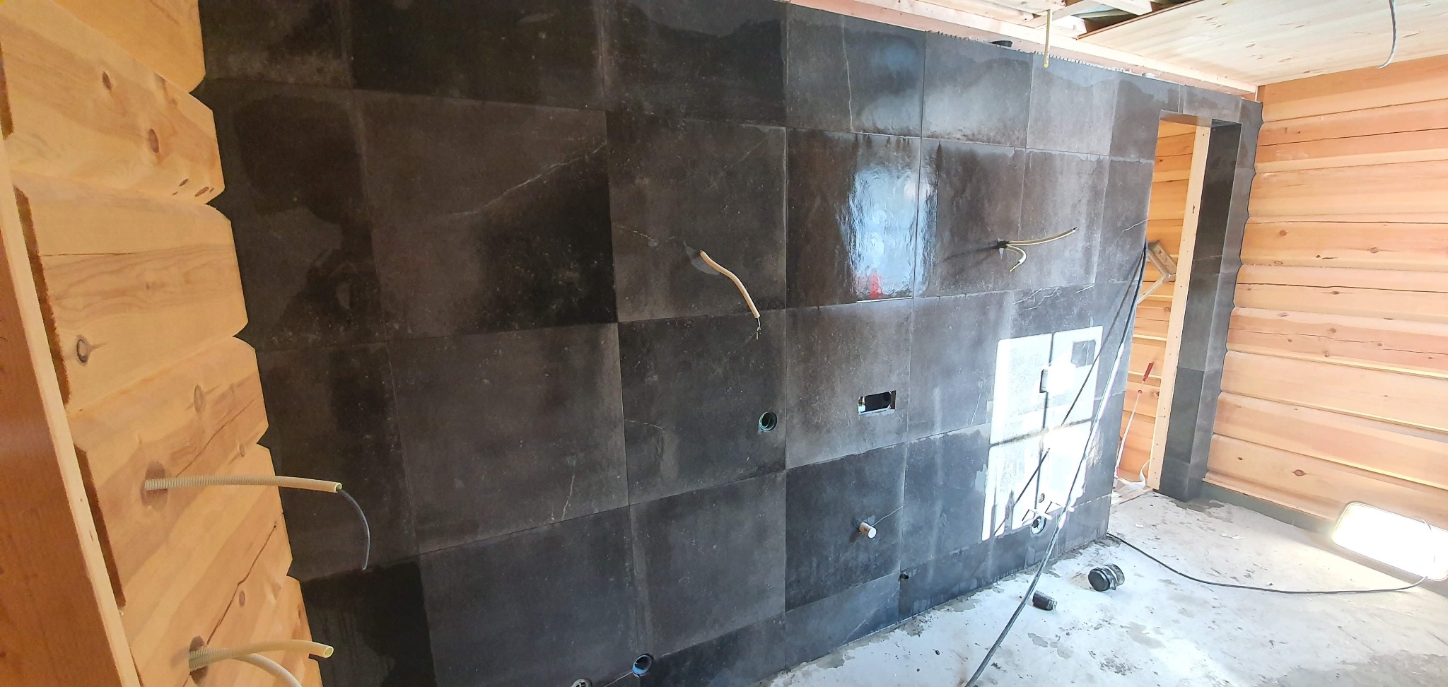 Ściana w łazience wyłożona ciemnymi, kwadratowymi płytkami z widocznymi instalacjami elektrycznymi i hydraulicznymi, w trakcie prac wykończeniowych w pomieszczeniu z drewnianymi ścianami.