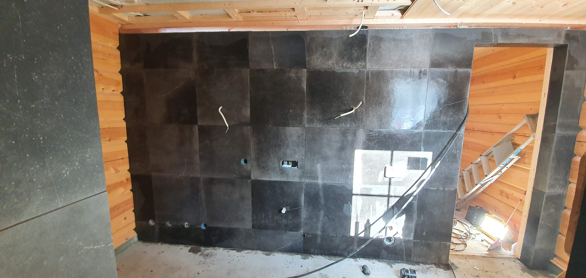 Ciemne płytki ułożone na ścianie w drewnianym domu, widoczne otwory na gniazdka i wystające przewody elektryczne, fragment drabiny i lampy oświetleniowej w przejściu.