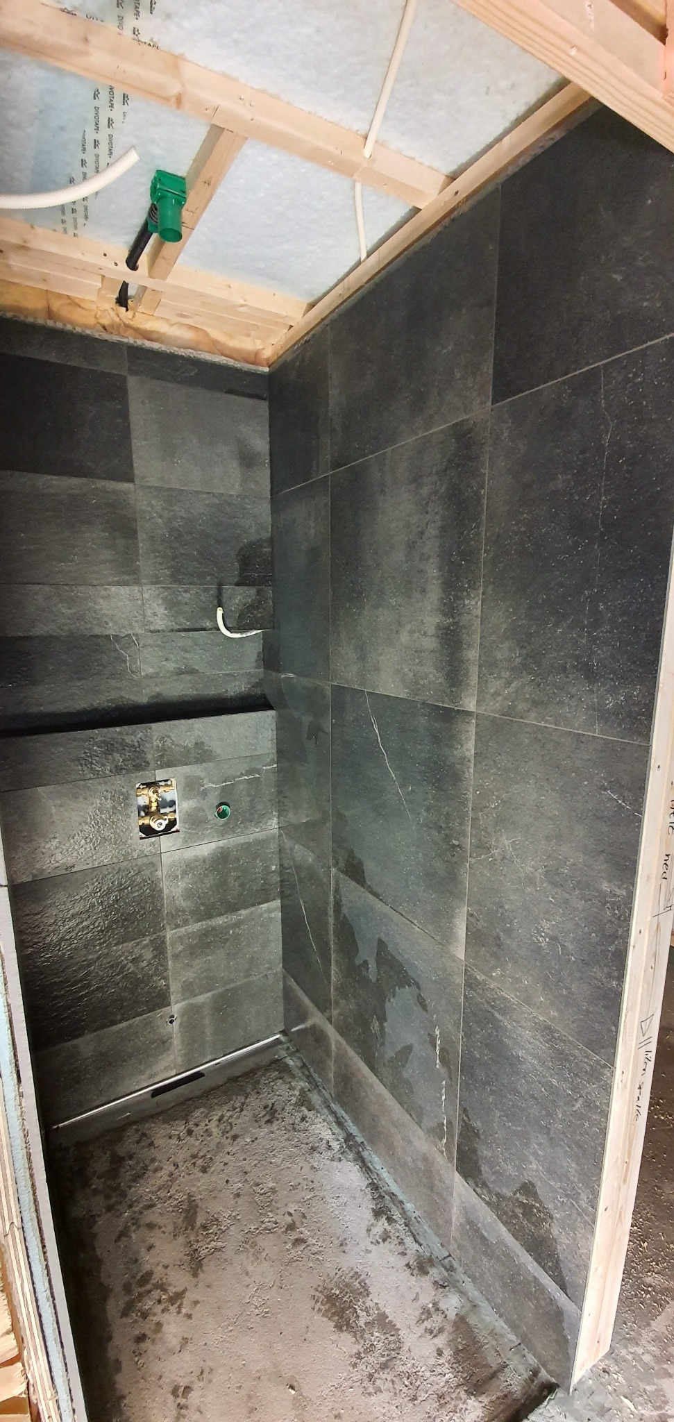 Wnętrze kabiny prysznicowej w trakcie prac wykończeniowych, widoczne ciemne płytki na ścianach, surowy beton na podłodze, instalacja wodna oraz drewniana konstrukcja sufitu i ścian.