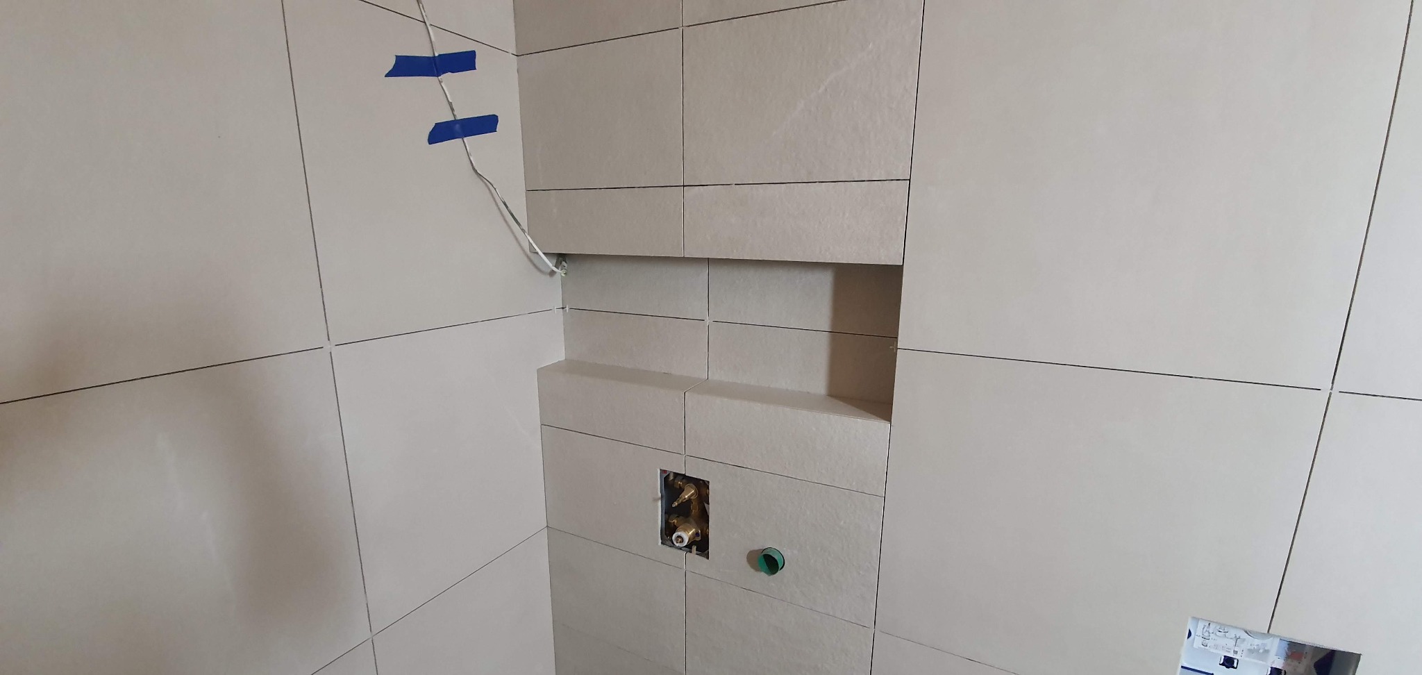 Ściana łazienki w trakcie kładzenia jasnych, kwadratowych płytek. Widoczna wnęka w ścianie oraz wystające przewody i rury z instalacji wodnej, zabezpieczone niebieską taśmą.
