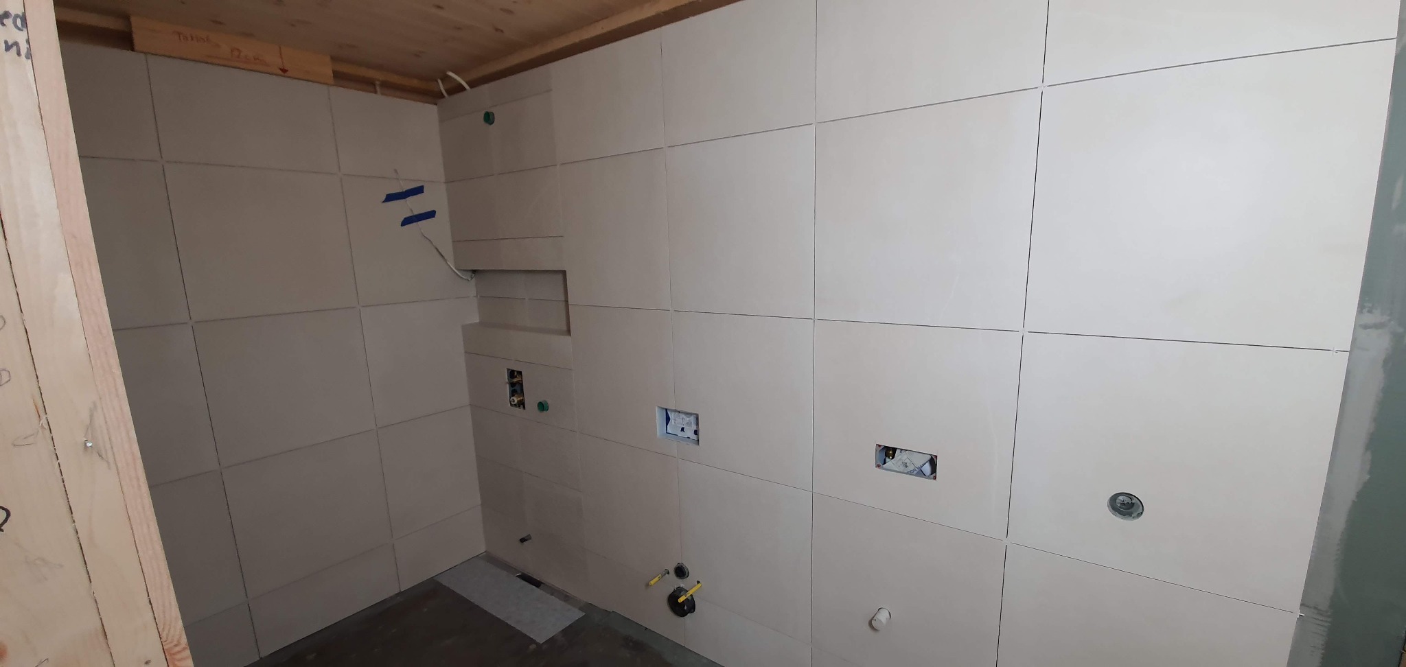 Łazienka w trakcie wykończenia: jasne, kwadratowe płytki na ścianach, widoczne otwory na instalacje elektryczne i hydrauliczne, niebieska taśma maskująca na górze, drewniane wykończenie sufitu.