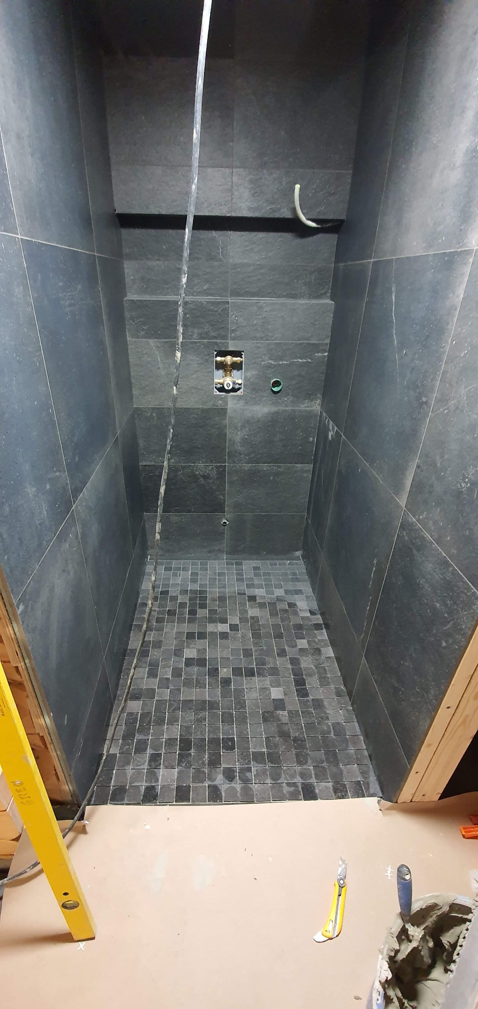 Montaż ciemnych płytek mozaikowych na podłodze w kabinie prysznicowej, widoczne narzędzia i poziomica, przygotowane do fugowania.