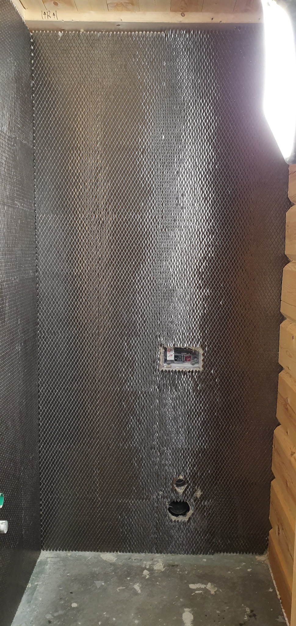 Przygotowana ściana w łazience z metalową siatką pod płytki, widoczne otwory na instalacje wodne i elektryczne, surowy beton na podłodze.