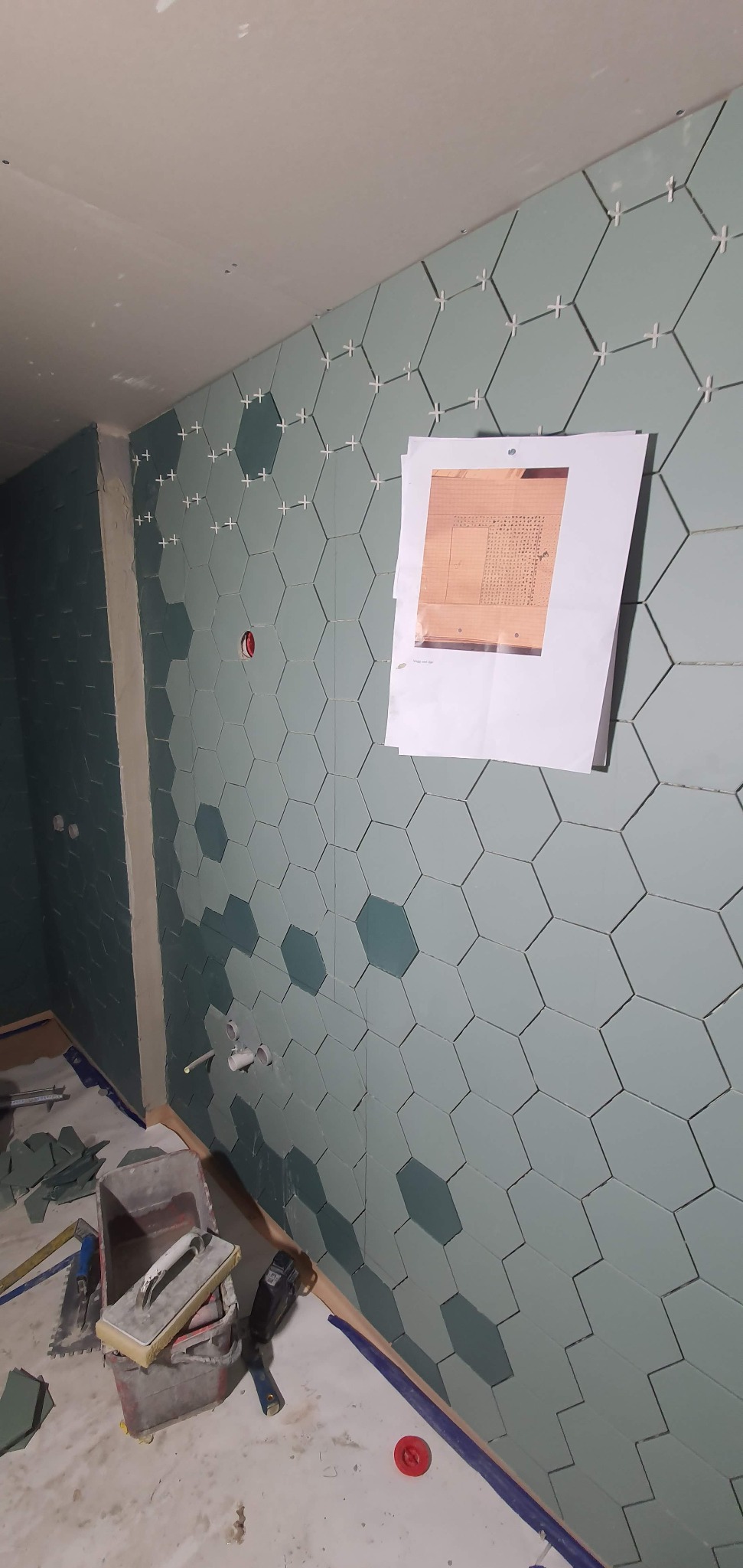 Ściana łazienki w trakcie układania heksagonalnych płytek w odcieniach zieleni, z widocznymi krzyżykami dystansowymi i planem ułożenia płytek przypiętym do ściany.