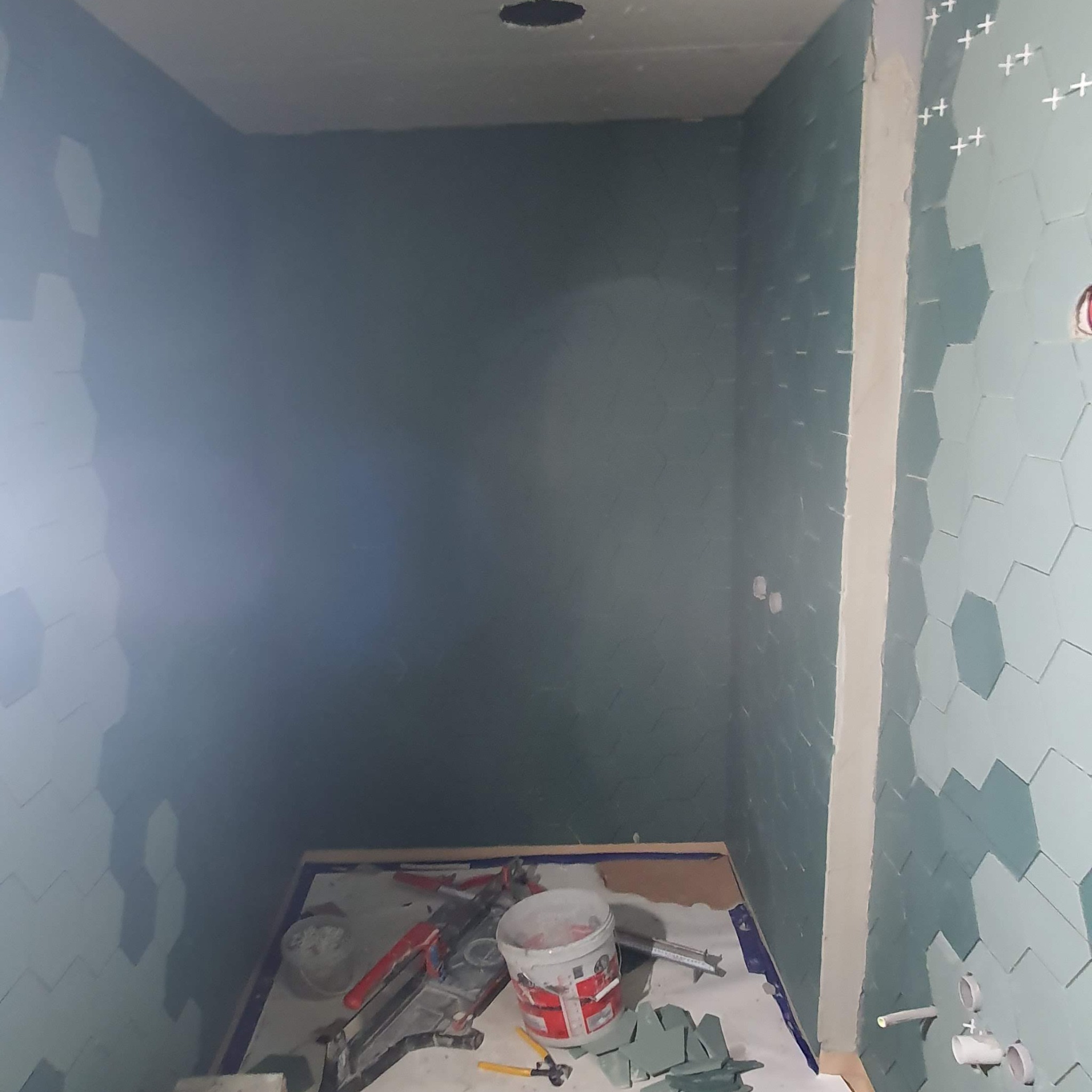 Wnętrze łazienki w trakcie układania heksagonalnych płytek w odcieniach szarości i błękitu, widoczne narzędzia glazurnicze, biała fuga i krzyżyki dystansowe.