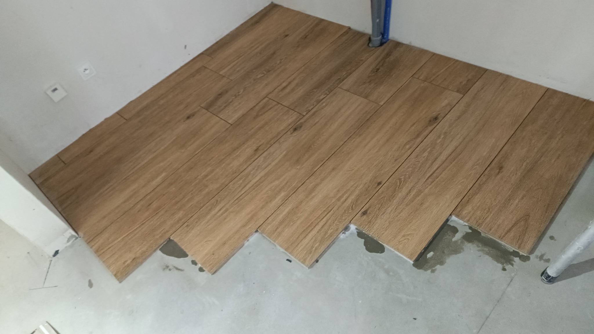 Układanie drewnopodobnych płytek na podłodze w rogu pomieszczenia, widoczne świeże ślady zaprawy.