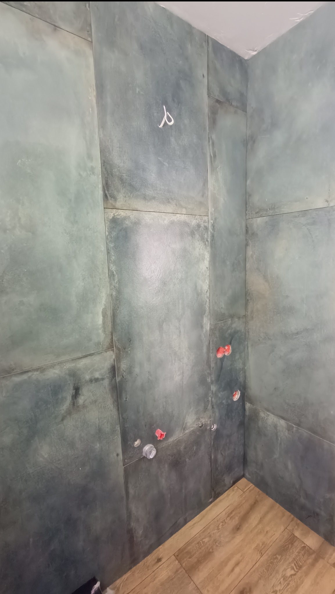 Ściany łazienki wyłożone dużymi, szarymi płytami imitującymi beton, z widocznymi wyprowadzeniami instalacji wodnej i fragmentem drewnopodobnej podłogi.