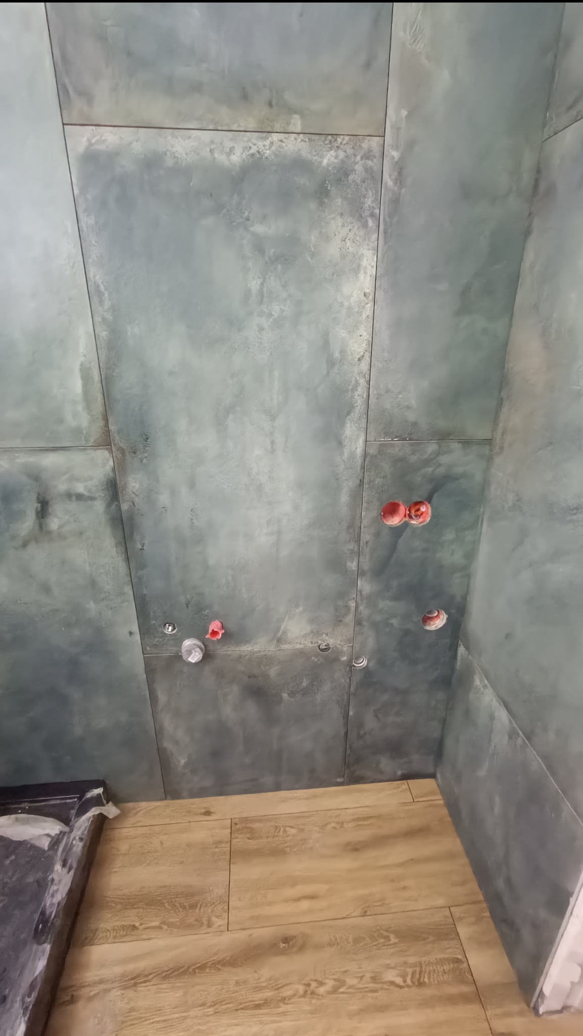 Ściana łazienki wyłożona szarymi płytami imitującymi beton, z widocznymi otworami na instalacje wodno-kanalizacyjne zabezpieczonymi czerwonymi i białymi zaślepkami, oraz podłoga wyłożona panelami...