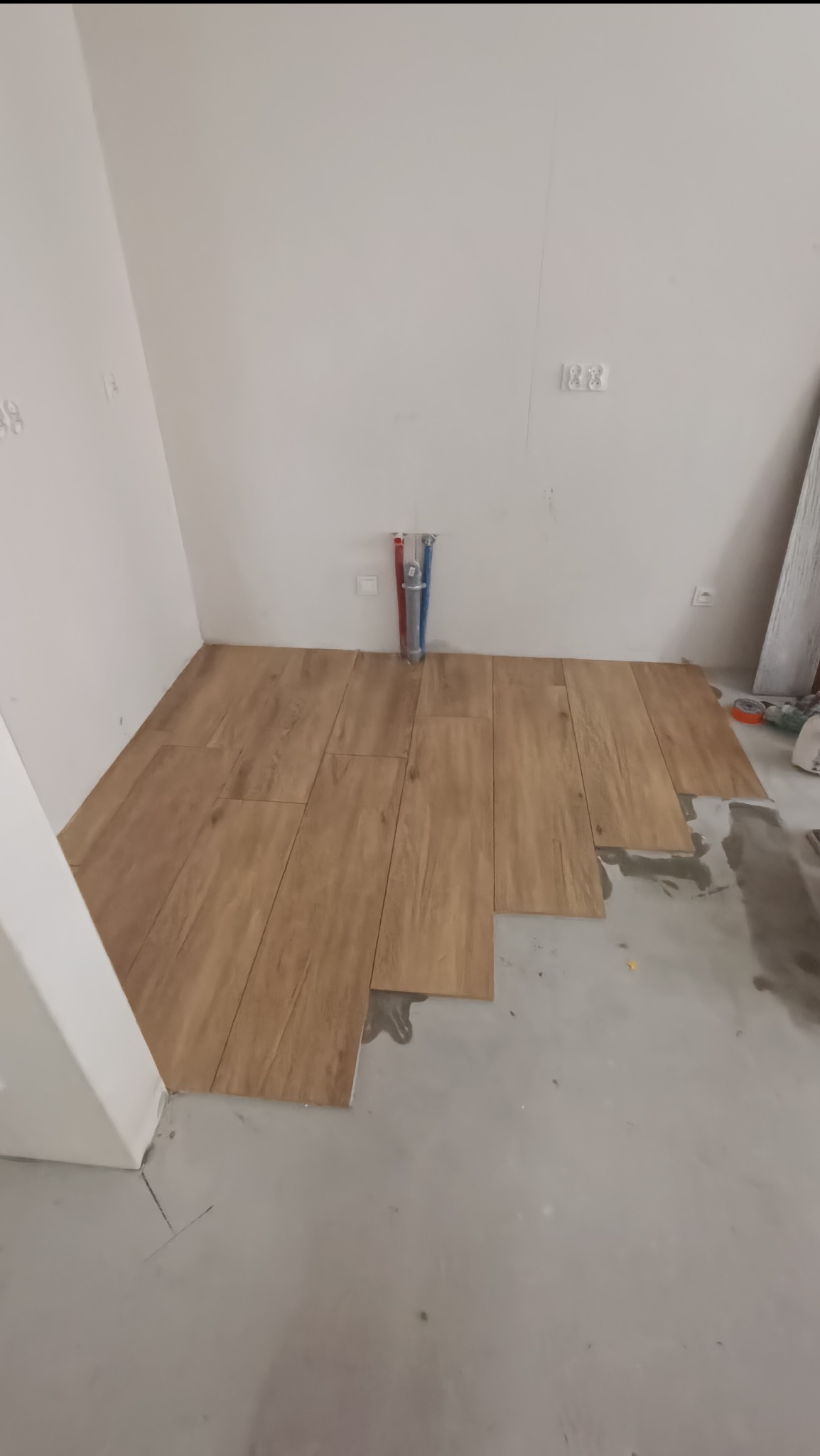 Układanie podłogi z płytek imitujących drewno w trakcie prac, widoczne rury instalacyjne na ścianie i częściowo położone płytki.