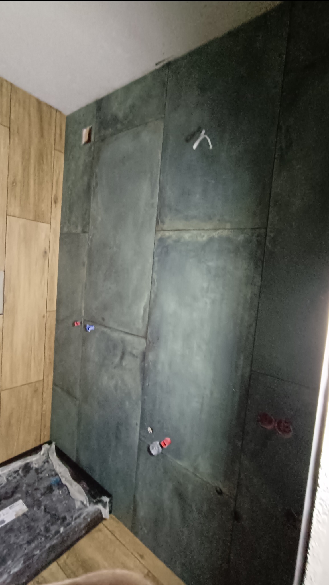 Ściana łazienki w trakcie układania ciemnych, kwadratowych płytek. Widoczne wystające rury instalacji wodnej i elektrycznej oraz plastikowe krzyżyki dystansowe pomiędzy płytkami.