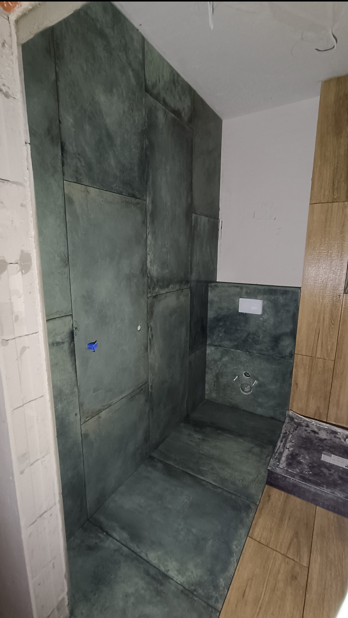Wnętrze kabiny prysznicowej w trakcie wykańczania, z ciemnymi płytkami imitującymi kamień na ścianach i podłodze, widoczne elementy instalacji wodnej i fragment drewnopodobnych płytek na podłodze.