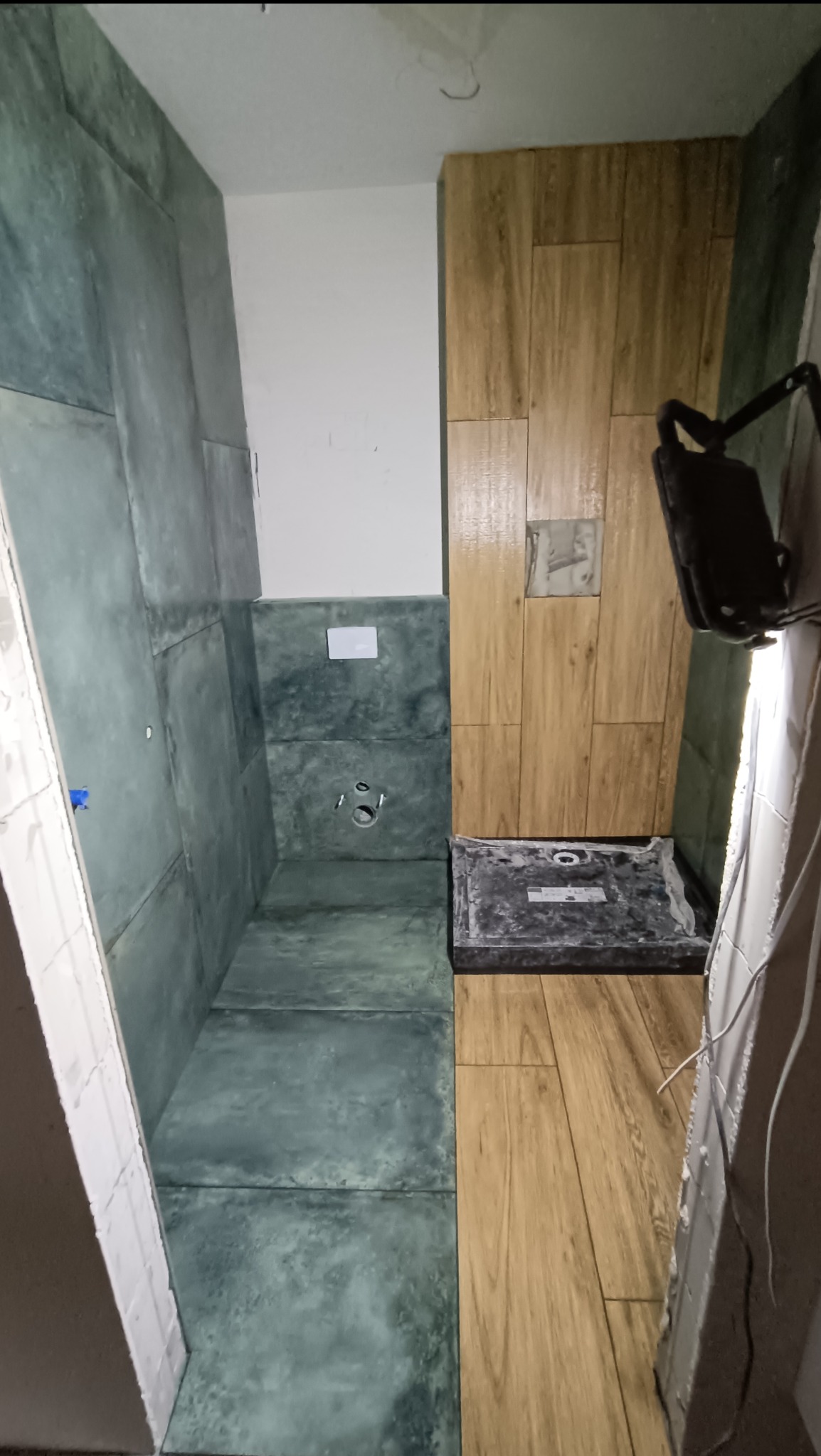 Wnętrze łazienki w trakcie prac wykończeniowych; widoczne płytki w odcieniach szarości i brązu, przygotowane otwory na armaturę, oraz przenośna lampa budowlana.