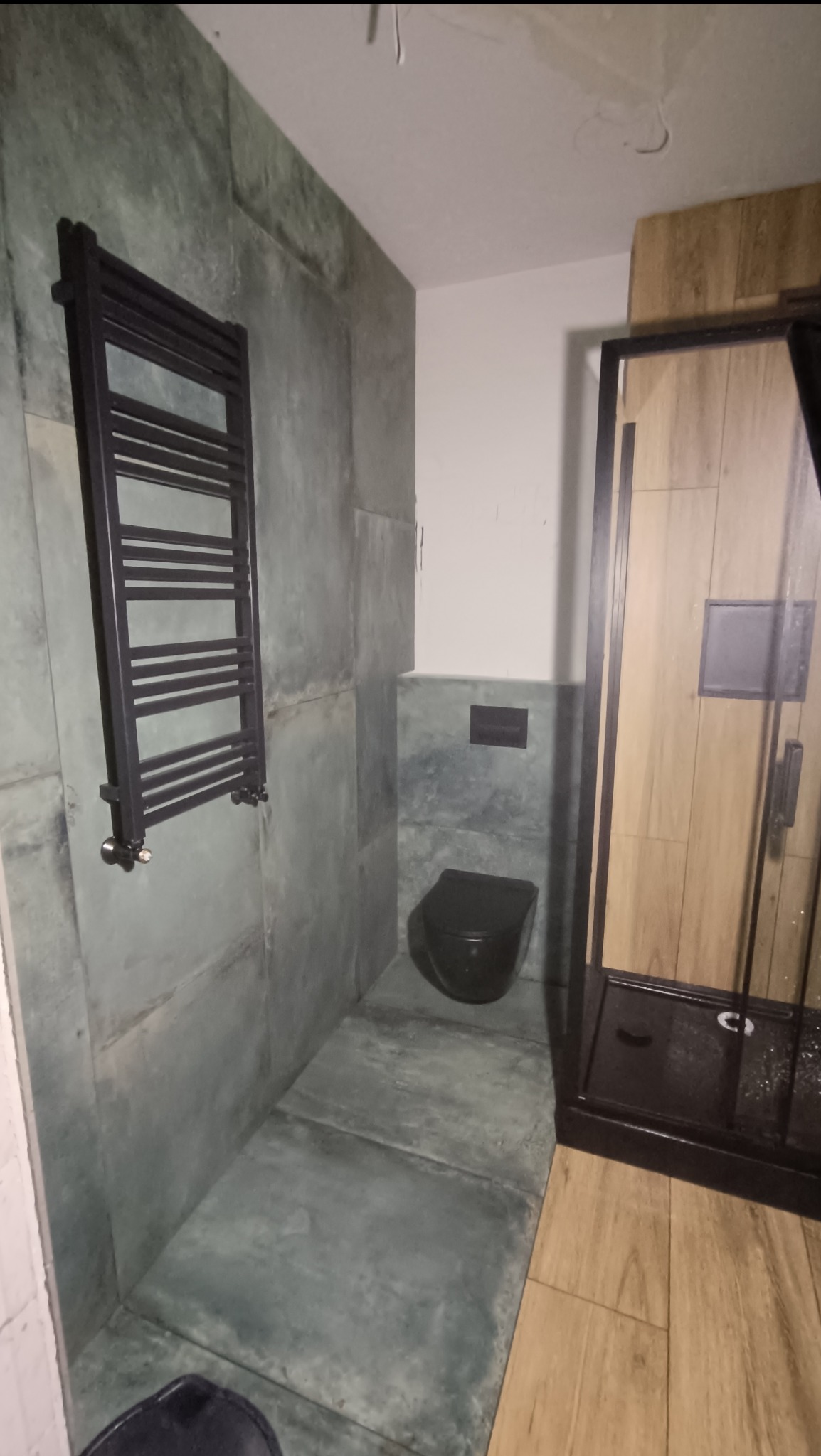 Wykończona łazienka w odcieniach szarości i drewna z wiszącym grzejnikiem drabinkowym, toaletą i kabiną prysznicową.