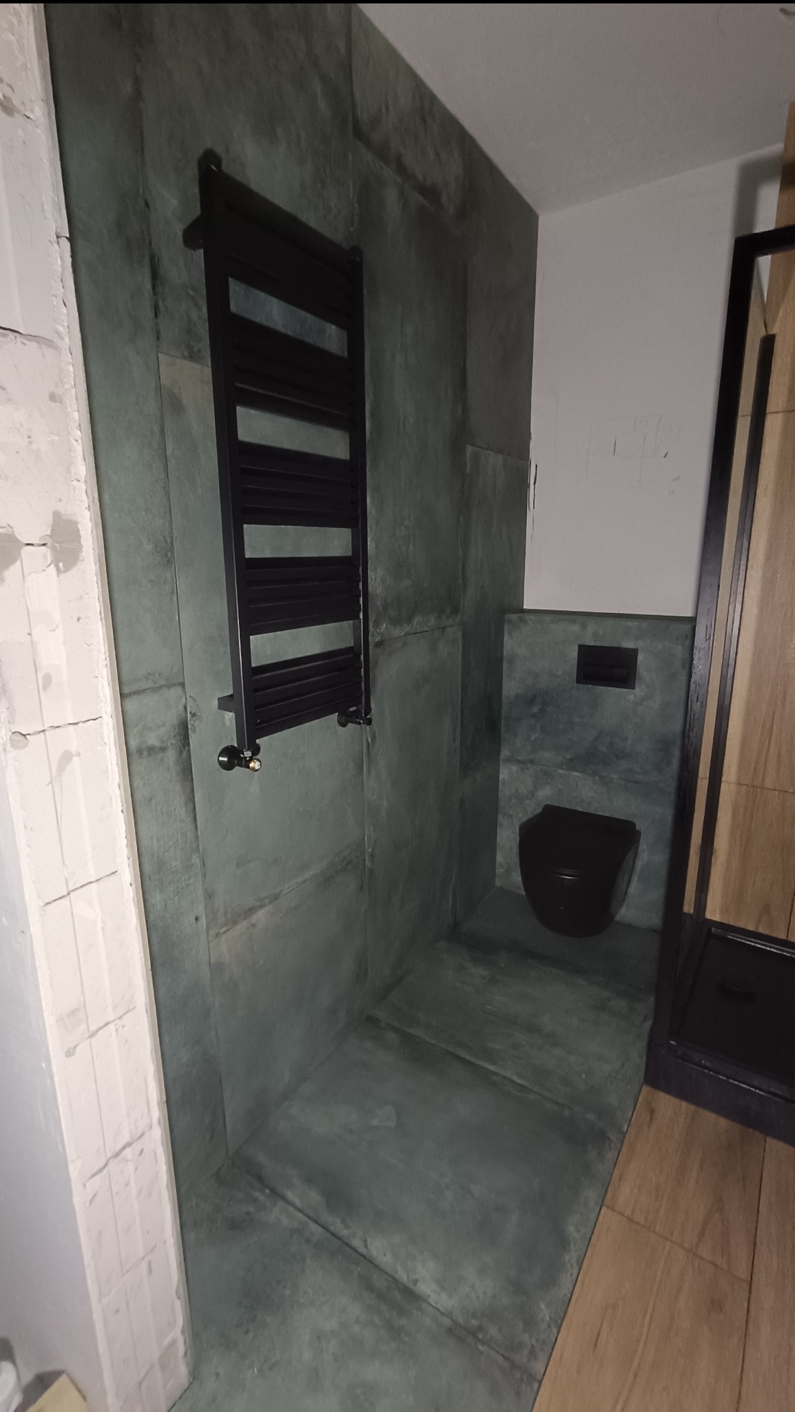 Nowoczesna łazienka z zielonymi, betonowymi płytami, czarnym grzejnikiem drabinkowym i podwieszaną toaletą.