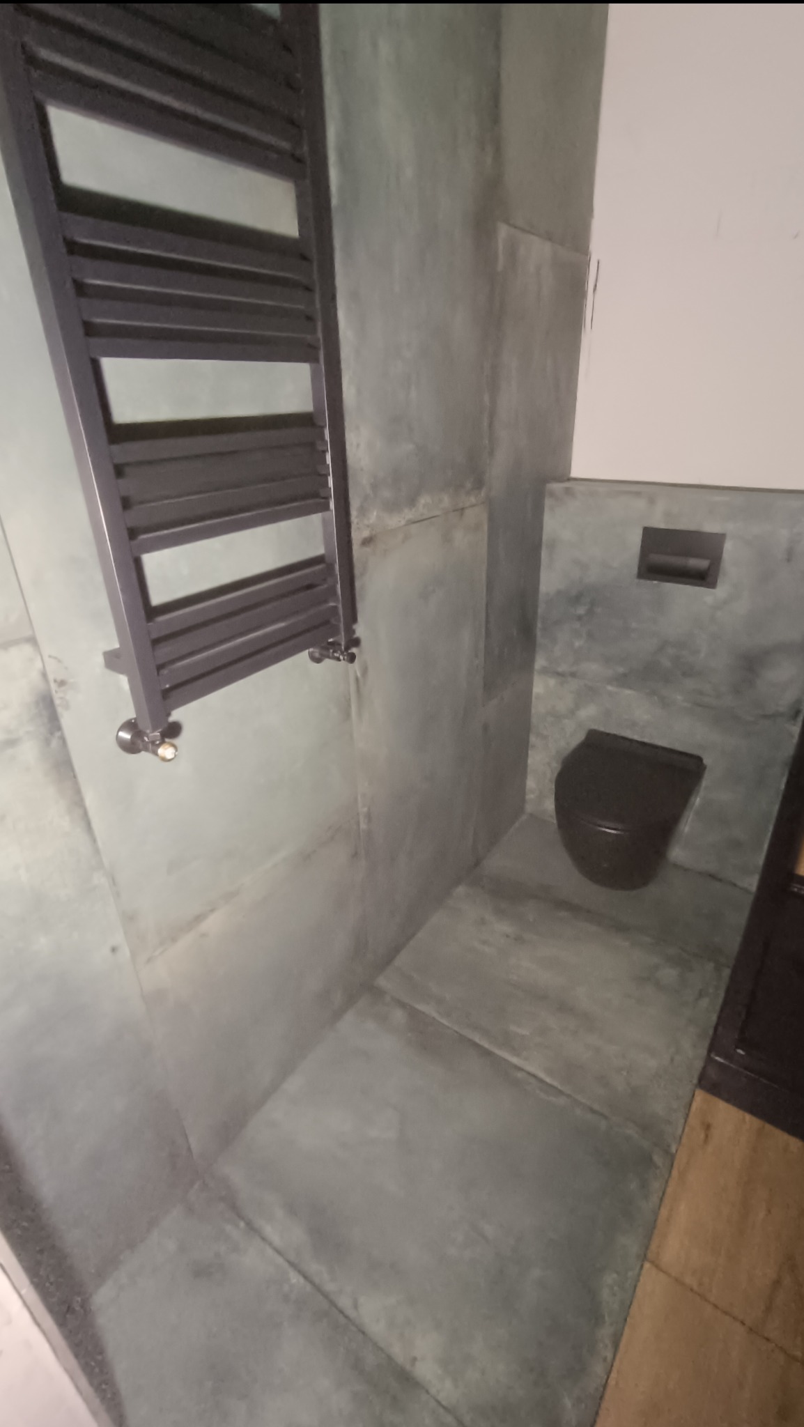 Nowoczesna łazienka z szarymi płytkami imitującymi beton, wiszącą toaletą w kolorze antracytowym i grzejnikiem drabinkowym w ciemnym kolorze, widok z perspektywy podłogi.