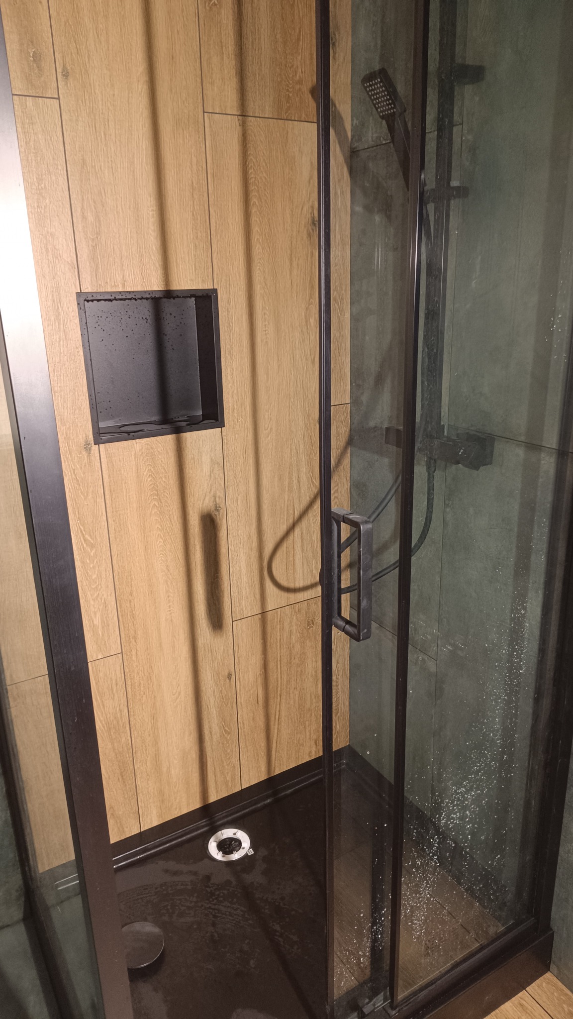 Kabina prysznicowa z czarnymi profilami i brodzikiem, drewnopodobne płytki na ścianie z wnęką.