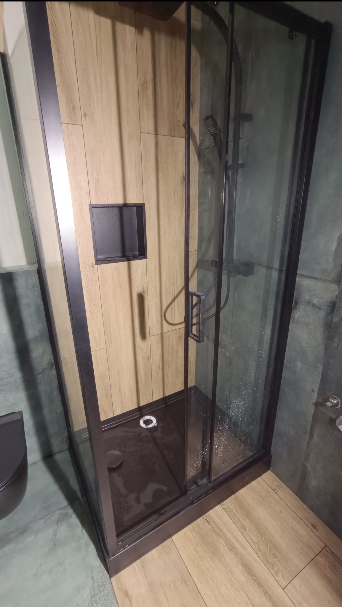 Kabina prysznicowa z czarną ramą i deszczownicą, obok toaleta w kolorze antracytowym, ściana wyłożona płytkami imitującymi drewno z wnęką na kosmetyki, widoczne ślady wody na szklanej powierzchni.