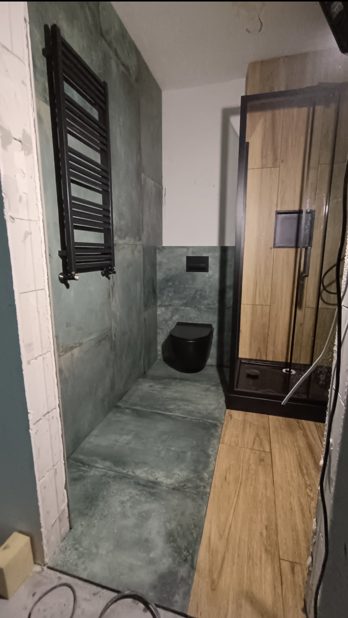 Wnętrze łazienki w trakcie prac wykończeniowych: ściany i podłoga wyłożone płytkami imitującymi beton, czarna toaleta podwieszana, grzejnik drabinkowy w kolorze czarnym oraz kabina prysznicowa...