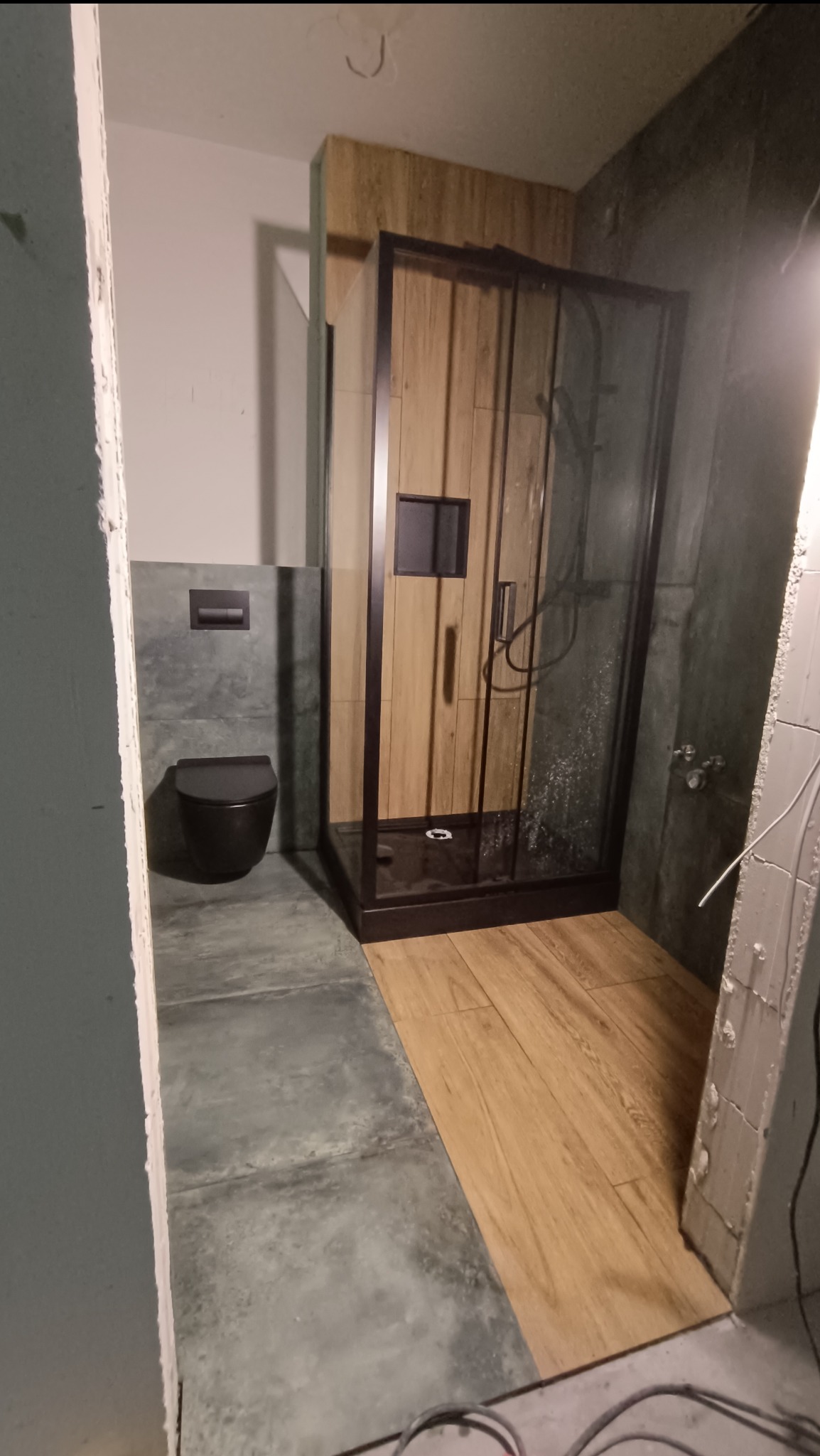 Częściowo wykończona łazienka z kabiną prysznicową z czarną ramą i drewnianymi panelami, wiszącą toaletą i szarymi płytkami podłogowymi, widoczne prace wykończeniowe przy ścianie.