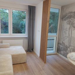 Halyna Khomanets - Jasne wnętrze po wykończeniu: drewniana podłoga, sofa, okno z widokiem na zieleń, dekoracyjna ścianka z drewnianych listew i tapeta w stylu retro z motywem drzew.