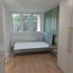 Halyna Khomanets - Jasne wnętrze sypialni po remoncie: drewniana podłoga, szara rama łóżka z zagłówkiem, białe ściany i szafa, akcentująca zielona ściana za łóżkiem, okno z widokiem na drzewa.