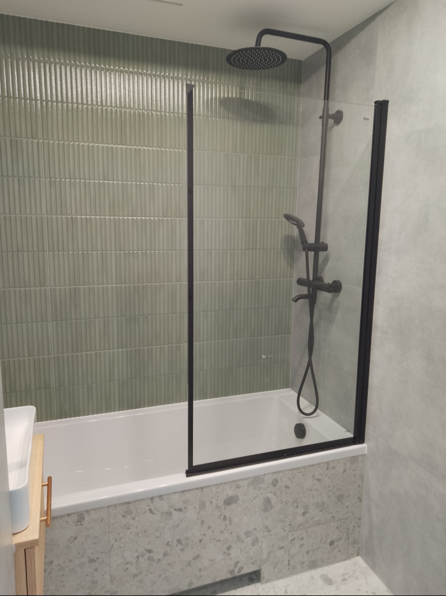 Nowoczesna łazienka: biała wanna z czarną armaturą, przeszklony parawan nawannowy z czarną ramą i zielone płytki na ścianie. Minimalistyczny design, jasne wnętrze.