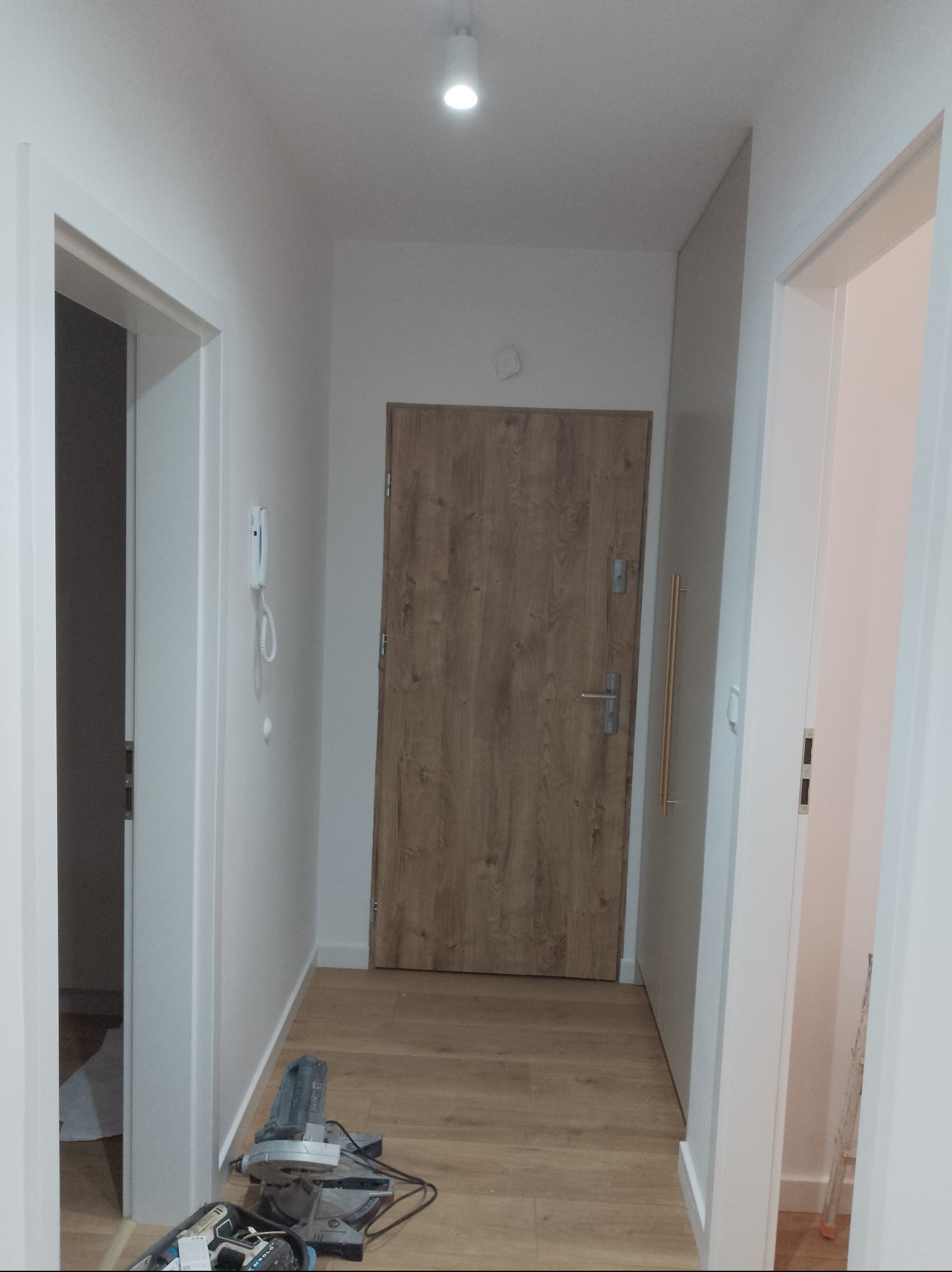 Jasne wnętrze po remoncie: drewnopodobne panele, białe ściany i stolarka. W tle drzwi wejściowe i narzędzia do montażu paneli.